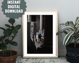 May include: A framed photograph of a person sitting on a chair in front of a large pillar in a building with a high ceiling. The person is wearing a dark jacket and a hat. The building has a white marble floor and a white marble column.