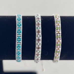 Könnte beinhalten: Drei Armbänder mit Perlen in Blau-, Grau- und Grüntönen. Jedes Armband besteht aus kleinen, farbigen Perlen und einer weißen Rückseite. Die Armbänder werden auf einer schwarzen Oberfläche präsentiert.