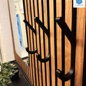 Könnte beinhalten: Eine Holz-Wandverkleidung mit vertikalen Holzlatten und schwarzen Metallhaken. Die Haken sind gebogen und an der Wand befestigt. Das Paneel ist Teil einer dekorativen Wand, möglicherweise für einen Flur oder Eingangsbereich.