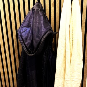 Auf dem Bild wird eine kleine Garderobe angezeigt, welche mit den Doppelhaken an ein Akustikpaneel-Wand bestückt sind. An den Haken hängen eine blaue Weste und ein weißer Schal.