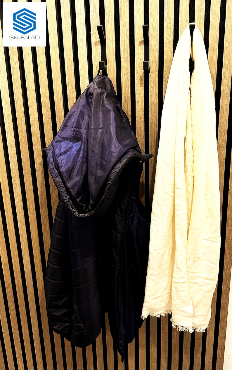 Auf dem Bild wird eine kleine Garderobe angezeigt, welche mit den Doppelhaken an ein Akustikpaneel-Wand bestückt sind. An den Haken hängen eine blaue Weste und ein weißer Schal.
