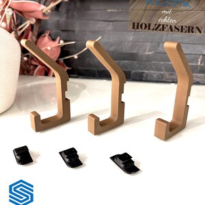 Auf dem Bild ist eine Einzelansicht von der Set-Option 3er-Set in Holzoptik aufgeführt. Es handelt sich um drei Doppelhaken in Holzoptik sowie drei Klemmsteine für die Montage ohne Bohren.
Auf dem Text oben rechts steht: "Enthält echte Holzfasern"