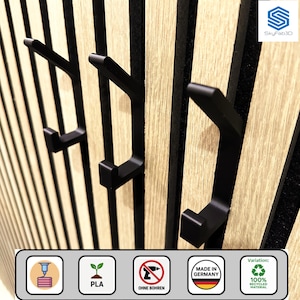 Könnte beinhalten: Schwarze Metall-Wandhaken, montiert auf einer Holzplatte mit vertikalen Streifen. Die Haken sind modern und minimalistisch gestaltet. Die Platte hat eine helle Holzmaserung. Das Bild enthält Symbole, die darauf hinweisen, dass das Produkt in Deutschland hergestellt wurde und aus recyceltem Material besteht.