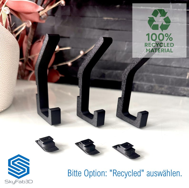 Auf dem Bild ist eine Einzelansicht von der Set-Option 3er-Set aufgeführt. Es handelt sich um drei schwarze Doppelhaken sowie drei Klemmsteinen für die Montage ohne Bohren.
Zusätzlich handelt es sich bei diesem Set um Haken aus 100% recyceltem Material. Als Text steht unten: "Bitte Option: "Recycled" auswählen".
