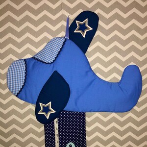May include: A blue airplane-shaped wall hanging with white star accents. The wings have a gingham pattern and a dark blue trim. A navy blue polka-dot fabric with a measuring tape and the name "AND" is attached to the bottom.