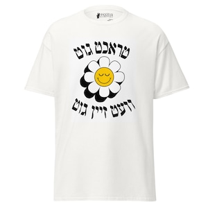 Può includere: T-shirt bianca con una grande grafica a margherita. La margherita ha un centro giallo con una faccia sorridente e petali neri e bianchi. La maglietta ha anche del testo in lingua straniera.