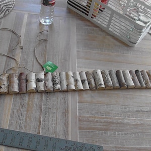 May include: A rustic wooden belt made of small, cylindrical pieces of birch bark. The belt is light brown and gray, with a natural, textured appearance. A string is attached to the belt, and a green tag hangs from the string. The belt is on a light brown wooden surface.