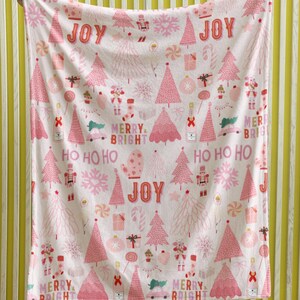 May include: A pink and white Christmas-themed blanket with a repeating pattern of Christmas trees, ornaments, and the words "JOY", "MERRY", and "BRIGHT". The blanket is held up by a person wearing black pants and white sneakers.