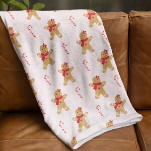 May include: A white blanket featuring a repeating pattern of gingerbread men with red scarves and candy canes. The blanket is draped over a brown leather sofa, creating a festive holiday theme.