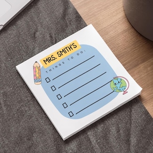 Puede incluir: Un bloc de notas blanco con un fondo azul y el texto "MRS. SMITH'S THINGS TO DO:". El bloc de notas presenta una ilustración de lápiz, un globo terráqueo y casillas de verificación para una lista de tareas. El bloc de notas está sobre una superficie gris.