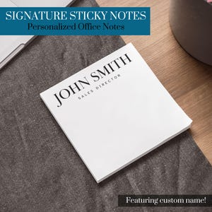 Könnte beinhalten: Weißer Haftnotizblock mit dem Text "JOHN SMITH SALES DIRECTOR" in Schwarz gedruckt. Der Block liegt auf einer grauen Stoffoberfläche.