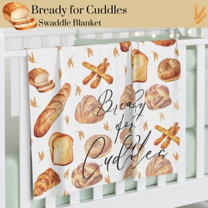 Pode incluir: Um cobertor de embrulho branco com um padrão de diferentes pães, incluindo baguetes, pães e pães. O texto "Bready for Cuddles" está escrito em caligrafia preta.