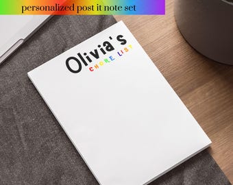 Puede incluir: Un bloc de notas blanco con el texto "Olivia's Chore List" en letras negras y arcoíris. El bloc de notas está sobre una superficie de tela gris.