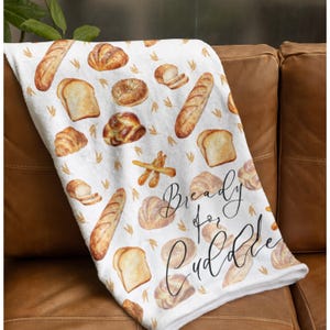 May include: White throw blanket with a pattern of various breads, including baguettes, loaves, rolls, and breadsticks. The text "Bready for Cuddle" is written in black cursive on the blanket.