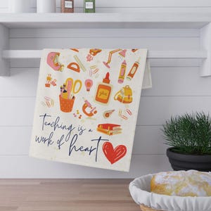 May include: A white kitchen towel with a colorful illustration of school supplies and the text "Teaching is a work of heart". The illustration includes a red heart, a pencil, a ruler, a pair of scissors, a glue bottle, a backpack, a microscope, a book, and a lamp.