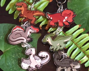 May include: Collection of dinosaur-themed keychains. Each keychain features a different cartoon dinosaur design in various colors, including red, brown, gray, and green. The keychains are attached to silver-colored metal clasps.
