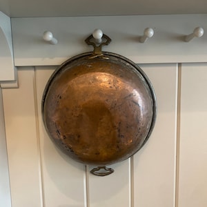 May include: A large, antique copper pan with a tarnished finish hangs on a white wall. The pan has a rounded shape with a metal rim and two small handles. Above the pan are four white, rounded hooks.