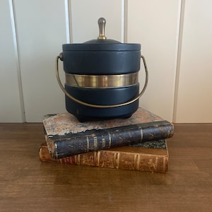 May include: A navy blue ice bucket with a gold-colored band and handle, topped with a lid and a small gold-colored handle. The ice bucket is sitting on top of two antique books with worn leather covers.