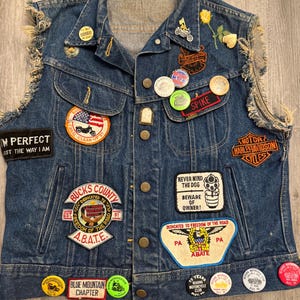 May include: A blue denim vest with frayed armholes, adorned with numerous embroidered patches and circular pinback buttons. Patches include Harley-Davidson logos, A.B.A.T.E. emblems, and text such as 'I'M PERFECT JUST THE WAY I AM' and 'NEVER MIND THE DOG'.