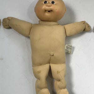 May include: A vintage Cabbage Patch Kid doll with a bald head, smiling face, and tan skin. The doll has outstretched arms and legs, with a soft body and a tag attached. The doll's features include painted eyes and a small, open mouth.