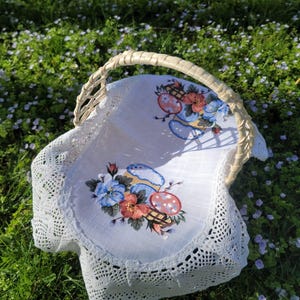 May include: A white wicker basket with a white linen liner featuring an embroidered design of blue and red flowers and mushrooms. The basket is sitting on a bed of green grass with small purple flowers.