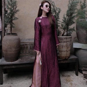 May include: A long, flowing burgundy dress with wide sleeves and a high slit, revealing a contrasting pink lining. The dress features floral embellishments near the neckline and is paired with pink shoes. The overall style suggests a formal or cultural garment.