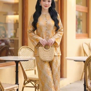 May include: A golden yellow two-piece outfit with a matching handbag. The long-sleeved top has a button-down front and the pants are wide-legged. The handbag is made of woven straw. The outfit is worn by a person standing in front of a cafe.