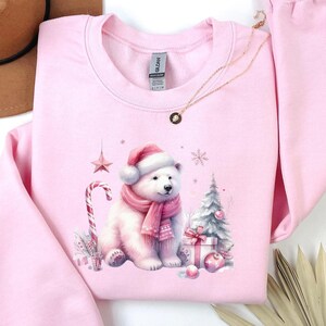 May include: Light pink sweatshirt with a Christmas-themed design featuring a polar bear wearing a Santa hat and scarf, holding a candy cane, and sitting next to a Christmas tree and presents.