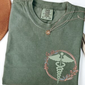 May include: A green t-shirt with a medical symbol design in a floral wreath. The symbol is green and the wreath is a light pink color. The t-shirt is folded and laid flat on a white surface.