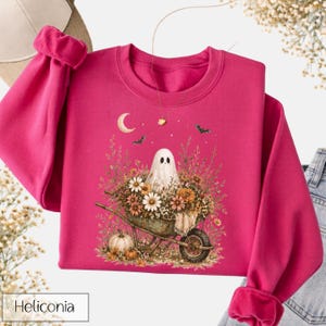 May include: A magenta sweatshirt featuring a whimsical Halloween design. The graphic includes a ghost in a wheelbarrow filled with flowers, a crescent moon, bats, and pumpkins. The word "Heliconia" is printed at the bottom.