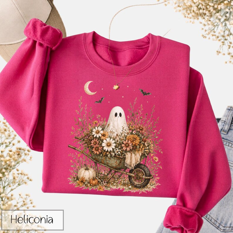 May include: A magenta sweatshirt featuring a whimsical Halloween design. The graphic includes a ghost in a wheelbarrow filled with flowers, a crescent moon, bats, and pumpkins. The word "Heliconia" is printed at the bottom.