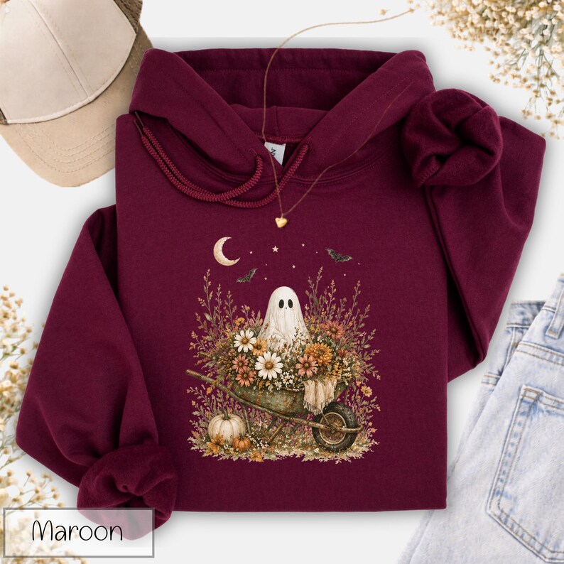 May include: Maroon hoodie with a whimsical ghost design. The graphic features a ghost in a wheelbarrow filled with flowers, a crescent moon, and bats. A gold necklace and a baseball cap are also visible.