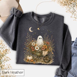 May include: Dark heather gray sweatshirt featuring a whimsical Halloween design. A white ghost is in a wheelbarrow filled with flowers and pumpkins. The design includes a crescent moon, stars, and bats. The text "Dark Heather" is at the bottom.
