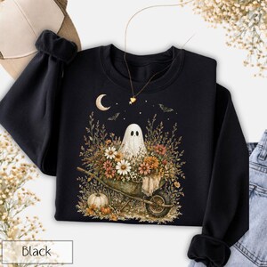 May include: Black sweatshirt featuring a whimsical Halloween design. The graphic depicts a friendly ghost in a wheelbarrow filled with flowers and pumpkins, under a crescent moon and bats. The word "Black" is printed at the bottom.