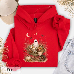 May include: A red hooded sweatshirt featuring a whimsical Halloween design. The graphic depicts a ghost in a wheelbarrow filled with flowers, a crescent moon, bats, and a pumpkin. The word "Red" is printed on the bottom left corner.