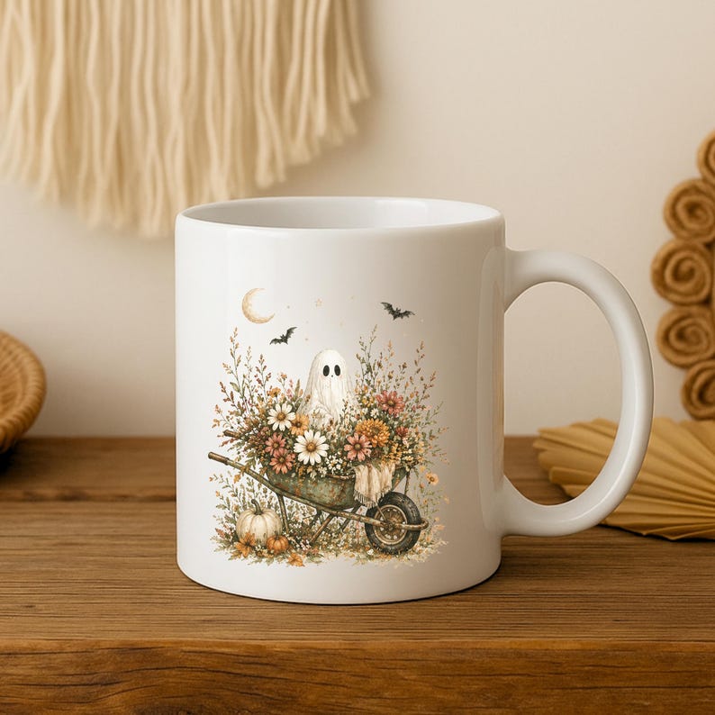 May include: White ceramic mug featuring a whimsical Halloween design. The illustration depicts a friendly ghost in a wheelbarrow filled with flowers and pumpkins. Bats and a crescent moon are also included in the design. The mug has a white handle and is set on a wooden surface.