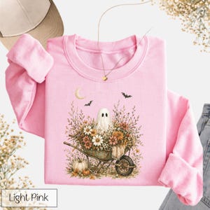 May include: Light pink crewneck sweatshirt featuring a whimsical illustration. The design includes a ghost in a wheelbarrow filled with flowers, a crescent moon, and bats. A pumpkin is also visible. The text "Light Pink" is in a white box at the bottom.