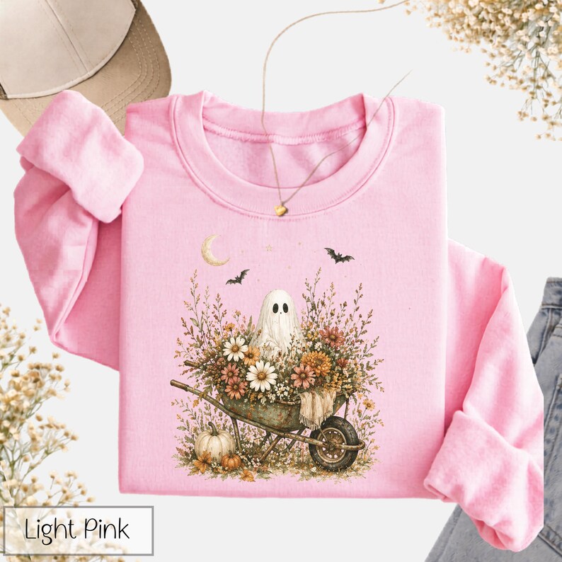 May include: Light pink crewneck sweatshirt featuring a whimsical illustration. The design includes a ghost in a wheelbarrow filled with flowers, a crescent moon, and bats. A pumpkin is also visible. The text "Light Pink" is in a white box at the bottom.