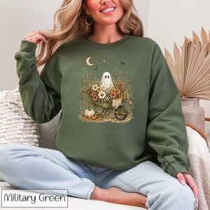 May include: Military green sweatshirt featuring a whimsical ghost design in a wheelbarrow filled with flowers, pumpkins, and a crescent moon. The design is printed on a comfortable crewneck sweatshirt.