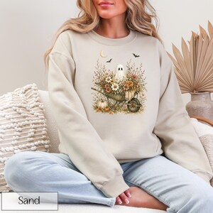 May include: Sand-colored sweatshirt featuring a watercolor design of a ghost in a wheelbarrow filled with flowers and pumpkins. The design includes a crescent moon and bats, perfect for Halloween or fall. The word "Sand" is printed at the bottom.