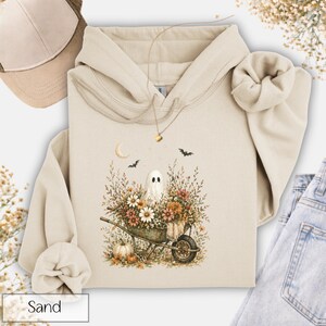 May include: Sand-colored hoodie featuring a whimsical Halloween design. The graphic depicts a ghost in a wheelbarrow filled with flowers, a crescent moon, and bats. A baseball cap is visible in the upper left corner. The word "Sand" is at the bottom.