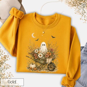 May include: Gold-colored sweatshirt featuring a whimsical Halloween design. The graphic includes a ghost in a wheelbarrow filled with flowers, a crescent moon, bats, and pumpkins. A gold necklace is visible. The word "Gold" is printed at the bottom left.
