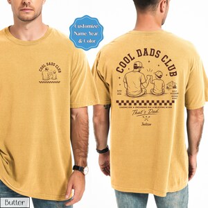 May include: Mustard yellow t-shirt with "COOL DADS CLUB" graphic on front and back. The back graphic features a father and child, mountains, and text including "That's Dad." The shirt includes the text "Customize: Name, Year, & Color."