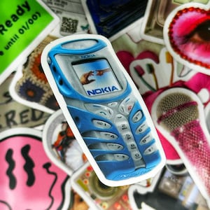 May include: A blue Nokia cell phone sticker with a white border. The phone has a digital display with a picture of two hands reaching towards each other. The phone has a keypad with the Nokia logo on the bottom.