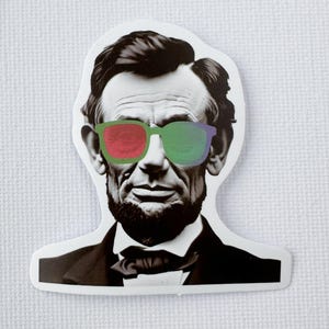 May include: A sticker depicting Abraham Lincoln in black and white, wearing sunglasses with red and green lenses. The sticker is cut to the shape of his head and shoulders.