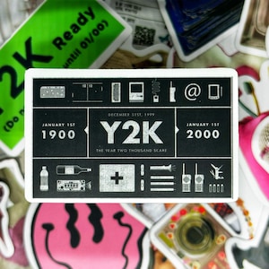 May include: A white sticker with black text and icons depicting the Y2K bug. The sticker reads "JANUARY 1ST 1900" and "JANUARY 1ST 2000" with "DECEMBER 31ST, 1999 Y2K THE YEAR TWO THOUSAND SCARE" in the centre. The icons include a computer monitor, a phone, an email symbol, a first aid kit, a set of three lines, a walkie talkie, and a padlock.