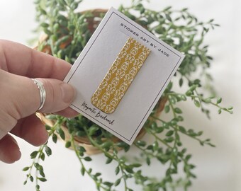 May include: A gold magnetic bookmark with a white patterned design. The bookmark is on a white card with the text "Sticker Art by Jade" and "Magnetic Bookmark".