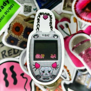 May include: A white and pink digital pet keychain with a pink heart charm and a silver chain. The screen displays a cartoon cat with a pink nose and ears. The text on the screen reads "Egg" and "Hungry".
