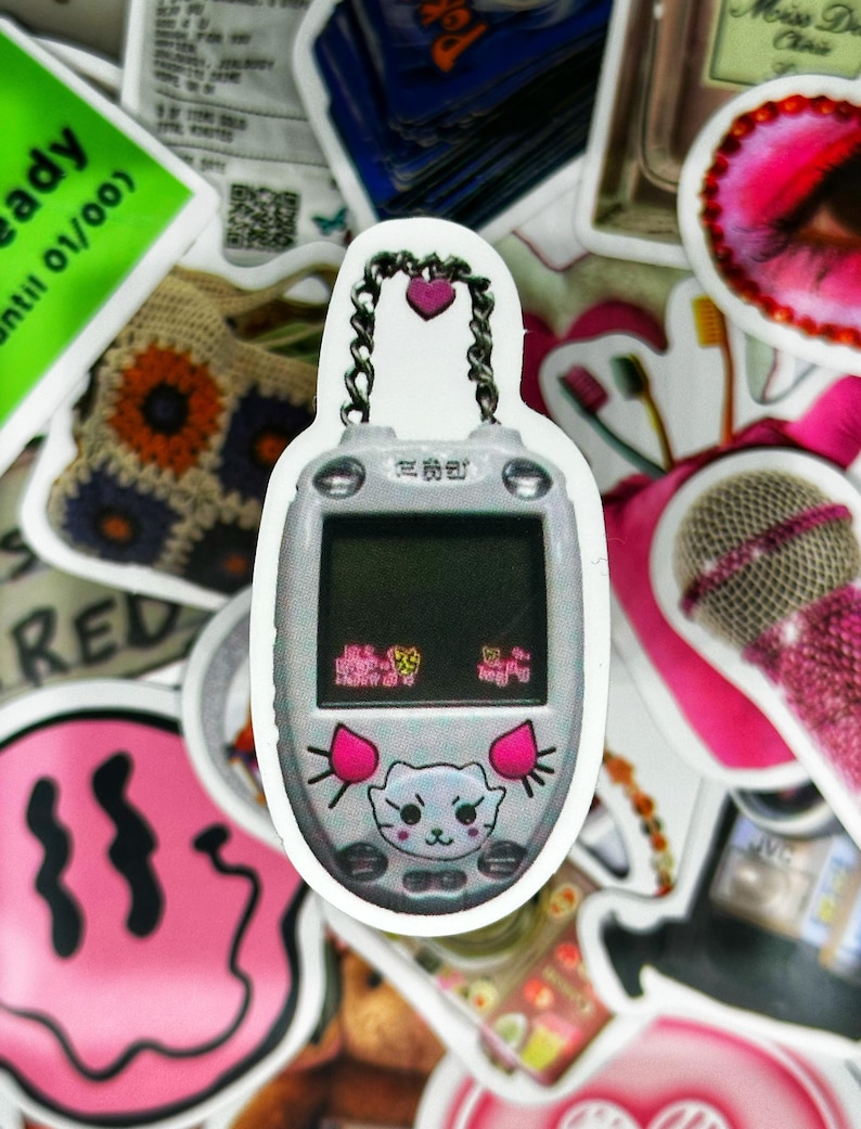 May include: A white and pink digital pet keychain with a pink heart charm and a silver chain. The screen displays a cartoon cat with a pink nose and ears. The text on the screen reads "Egg" and "Hungry".