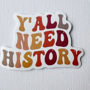 May include: A white sticker with the text "Y'ALL NEED HISTORY" in a retro font. The text is in shades of brown, orange, and red. The sticker has a white border and is on a textured white background.